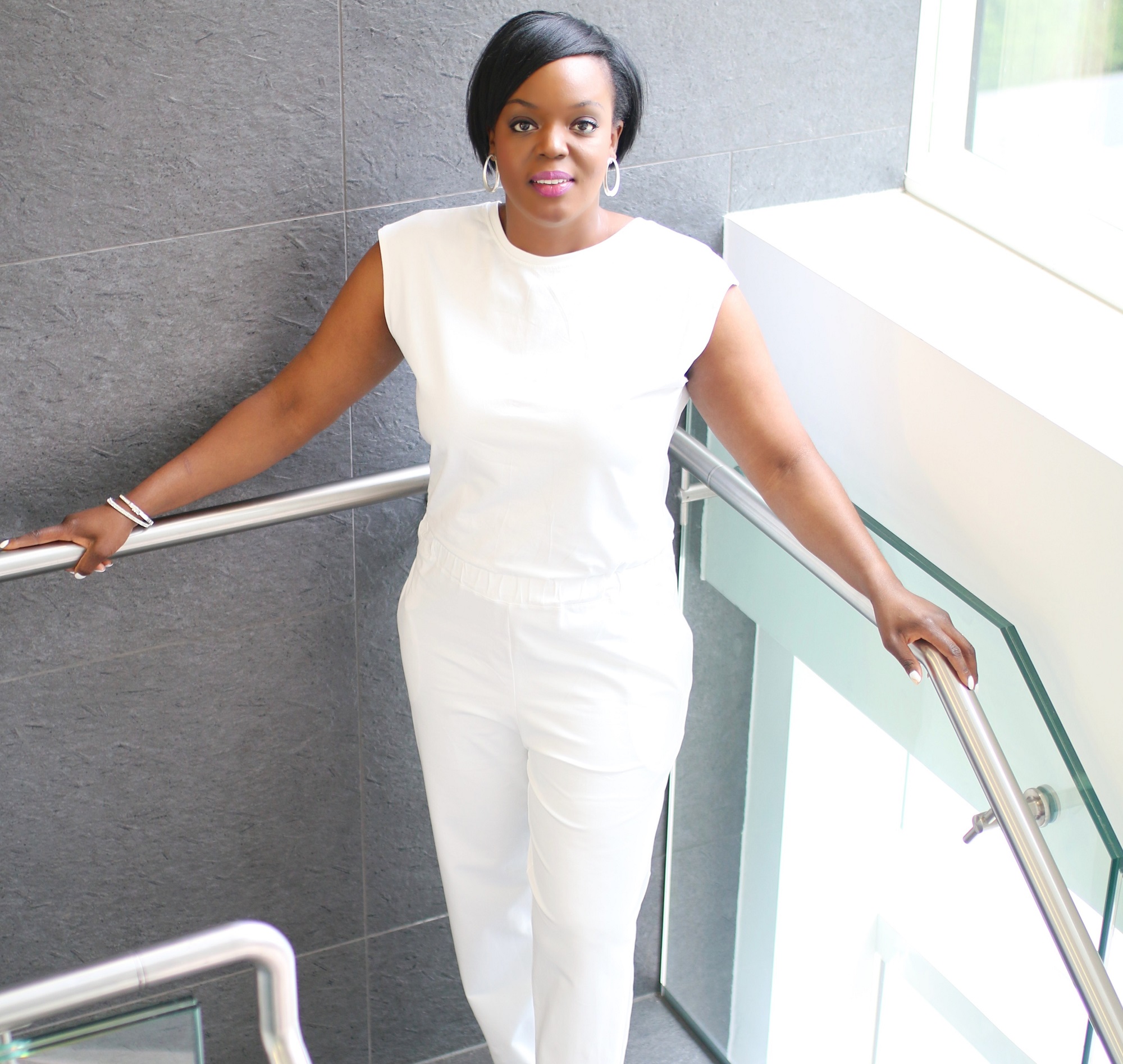 A woman in a white sleeveless jumpsuit with a ruched waist holding onto metal railings on a modern staircase.