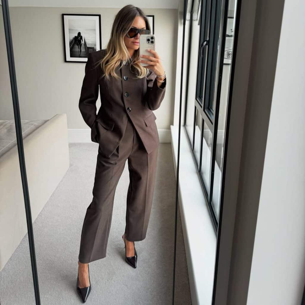 A woman with blonde hair taking a mirror selfie in a tailored brown suit with black pointed heels.