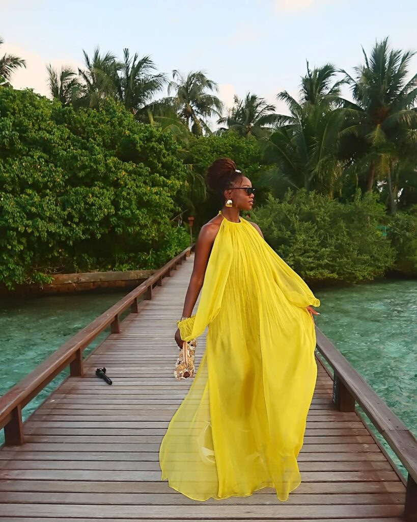 A woman in a vibrant yellow halter-neck maxi dress walking along a wooden boardwalk over turquoise water.
