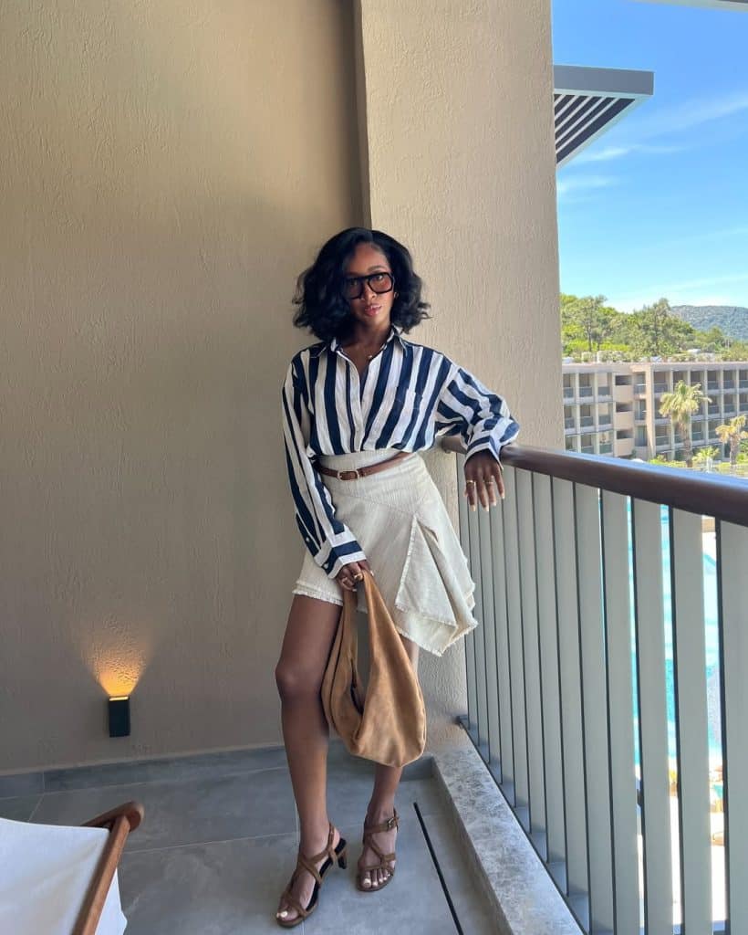 A woman in a striped shirt and cream asymmetrical skirt posing on a sunny balcony.