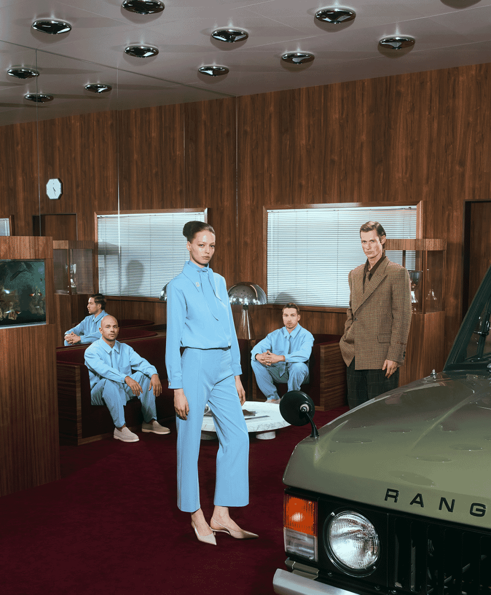 Models in light blue tailored suits in a retro-futuristic wood-paneled room with a vintage car.