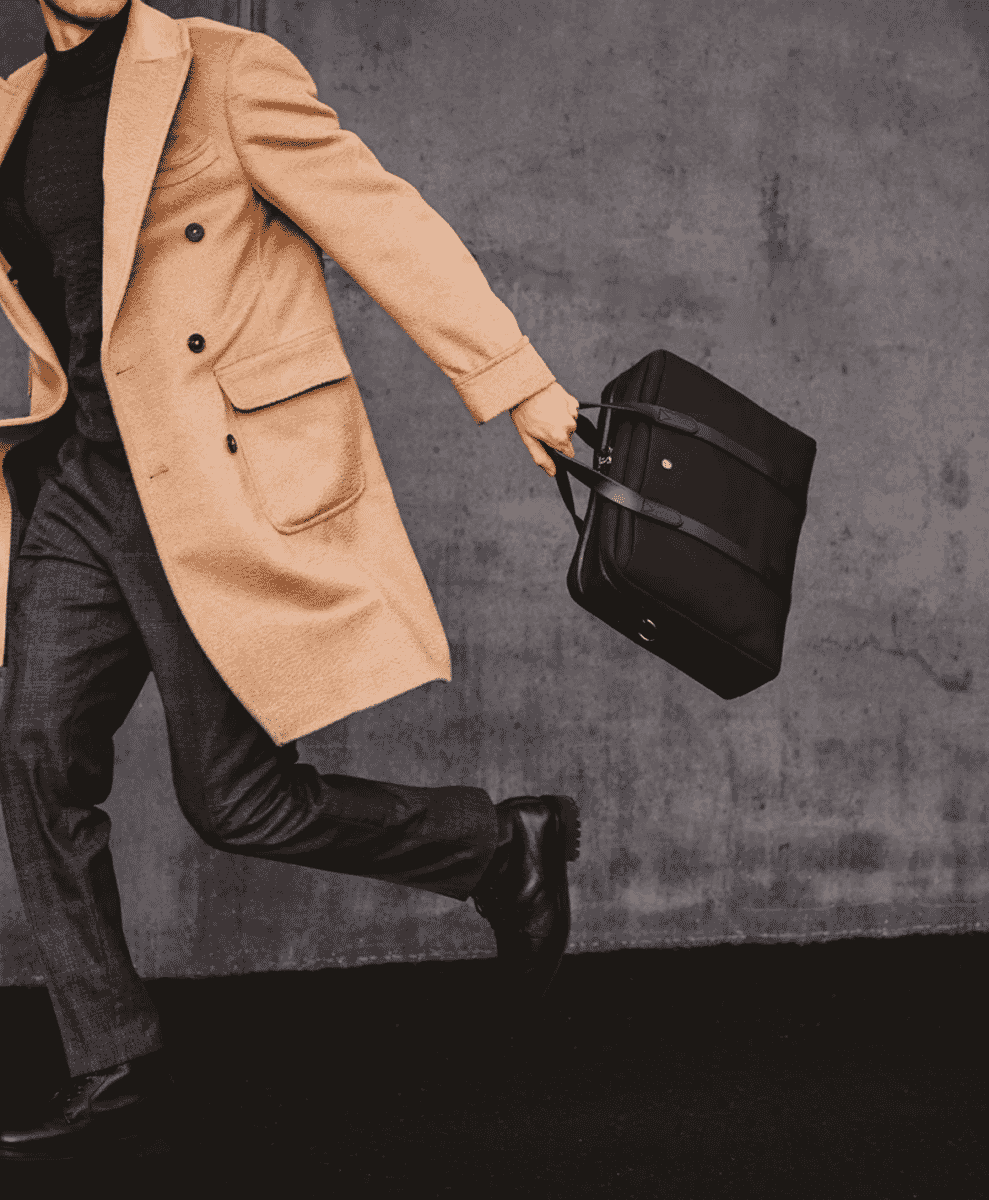 A man in a beige wool overcoat and black turtleneck carrying a briefcase.