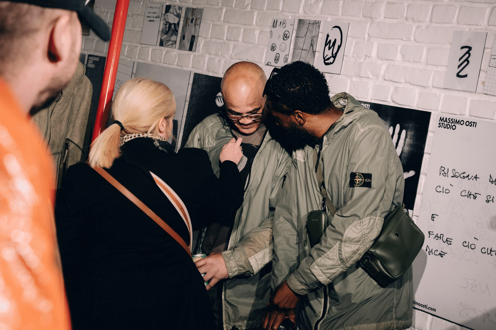 Three people in technical streetwear gathered near a white brick wall with fashion sketches and studio branding.