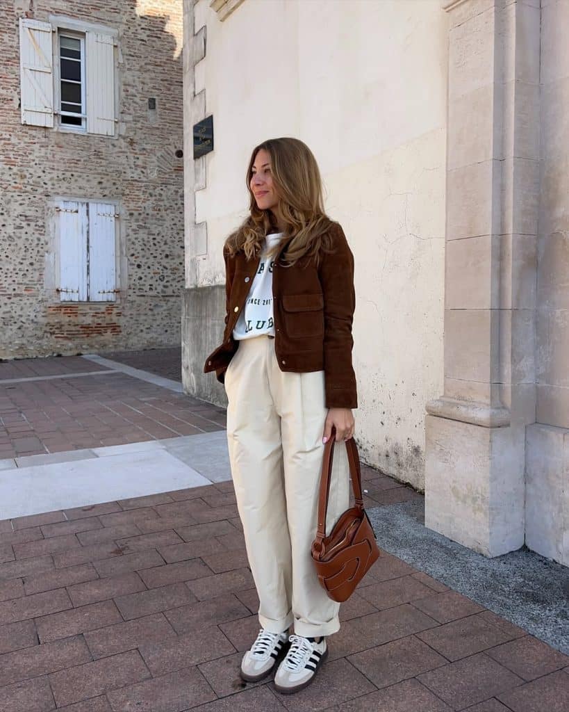 A woman in a brown suede jacket and cream wide-leg pants walking on a cobblestone street.