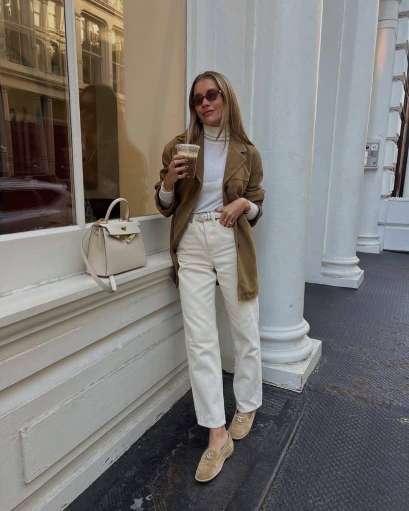 A woman in a camel coat, white turtleneck, and cream wide-leg jeans standing in an urban setting.