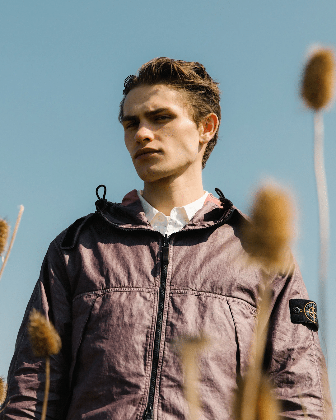 A man wearing a mauve textured Stone Island hooded jacket against a blue sky.