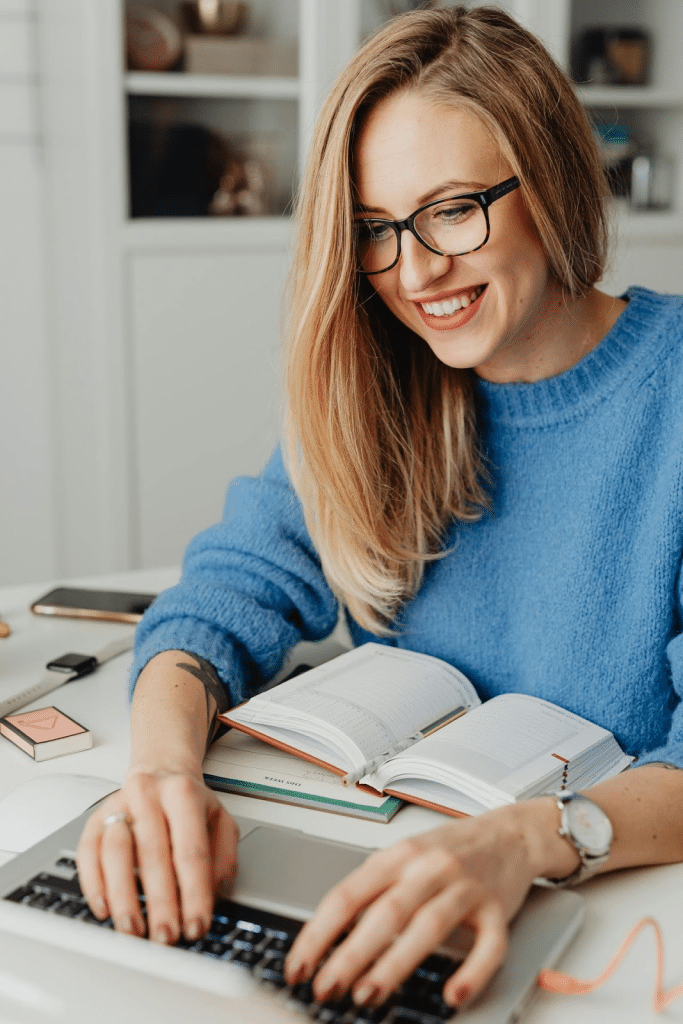 A professional woman wearing a blue knit sweater and black glasses working on a laptop at a desk.