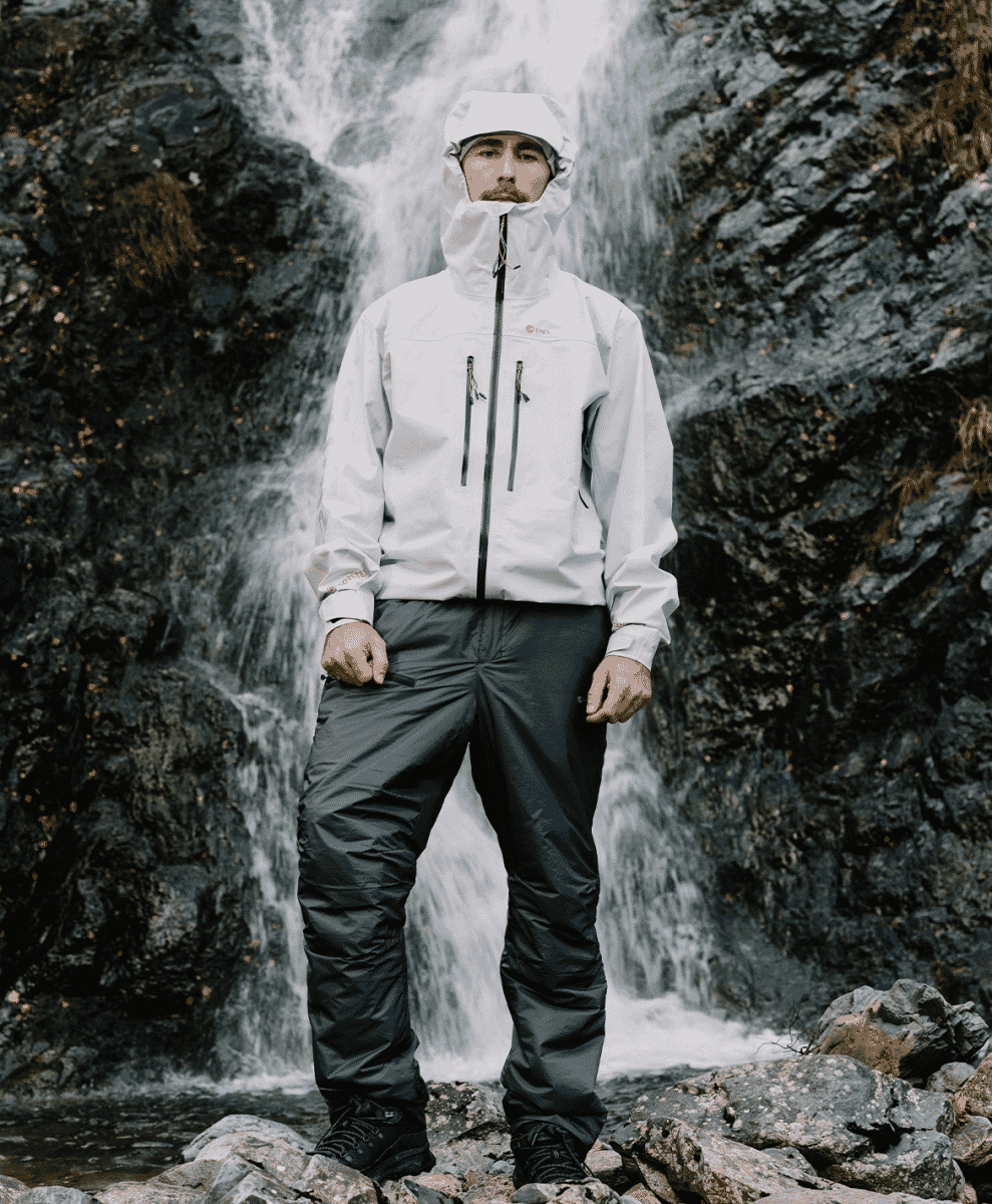 A man in a white hooded waterproof jacket standing before a large, misty waterfall.