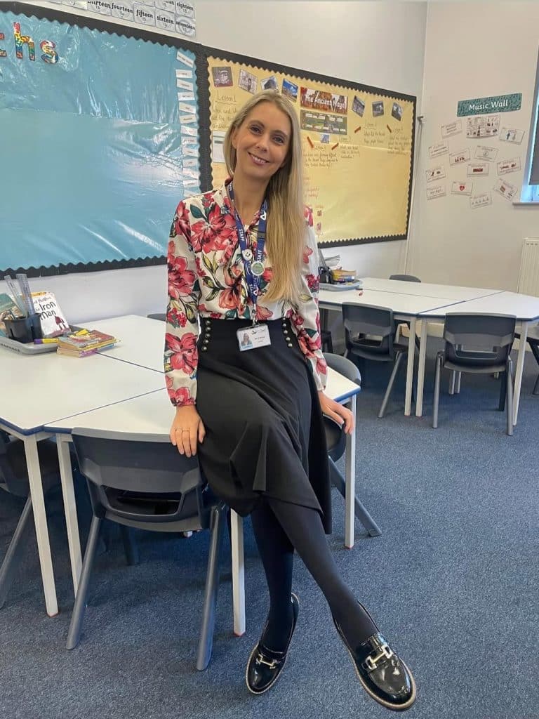 A woman wearing a floral blouse and patent leather loafers in a classroom.