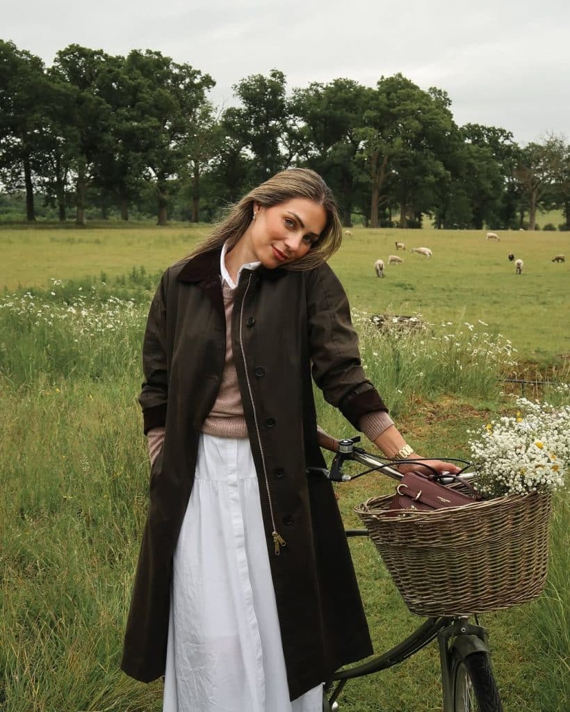 A woman in a brown waxed coat and white skirt posing with a bicycle in a pastoral field.