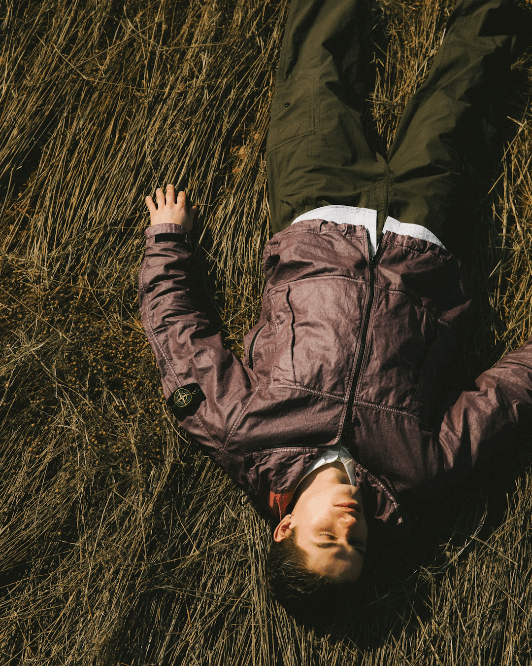 Aerial shot of a man lying in dry grass wearing a textured brown Stone Island jacket and olive cargo pants.