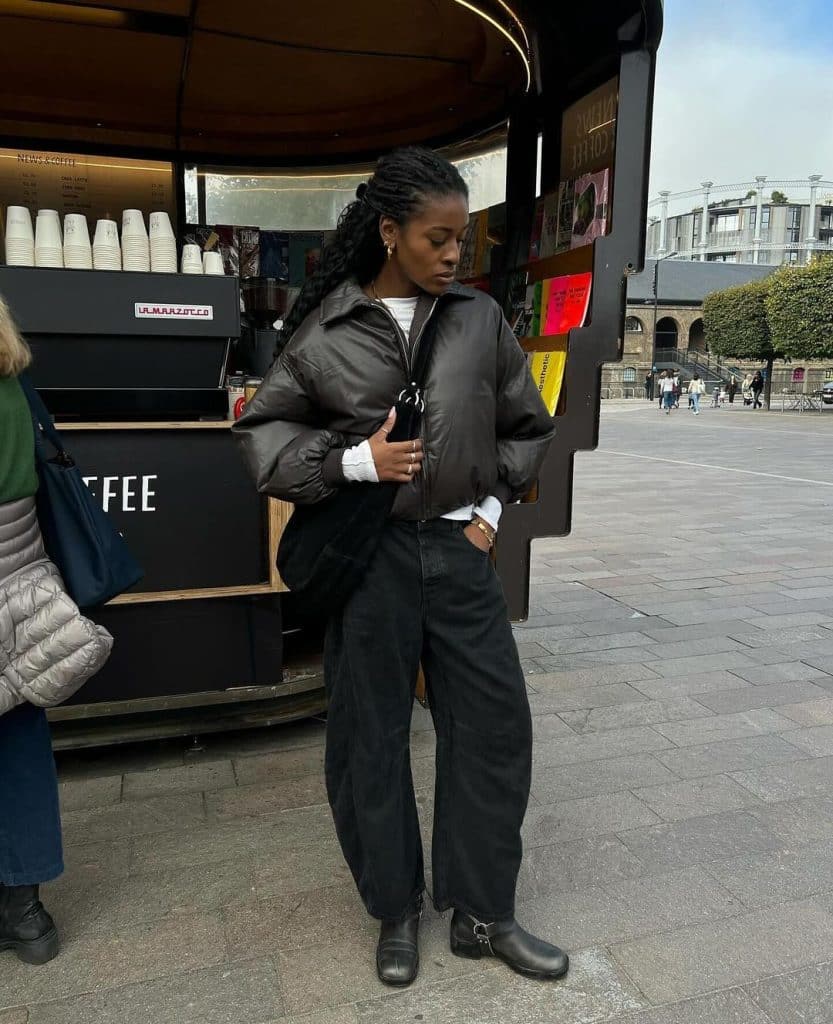 Woman in a dark brown puffer jacket and black barrel-cut trousers standing near a coffee cart.