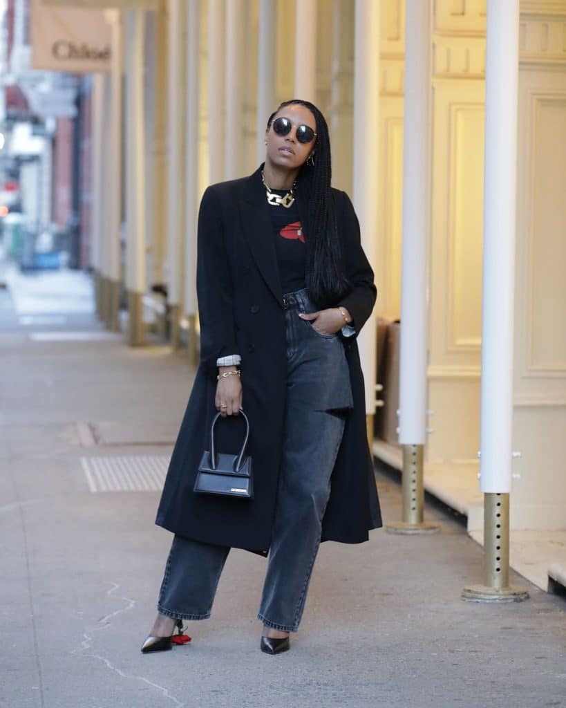 Woman walking on a city sidewalk wearing a black double-breasted wool coat and blue barrel jeans.