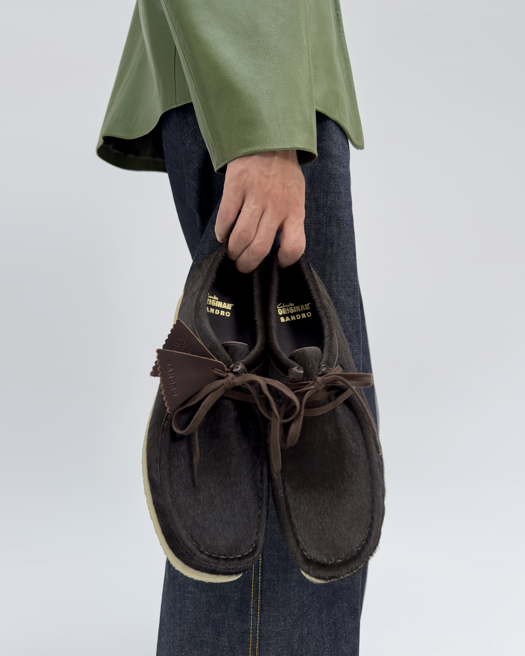 A person in a green leather jacket holding the brown Clarks Originals Sandro moccasins.
