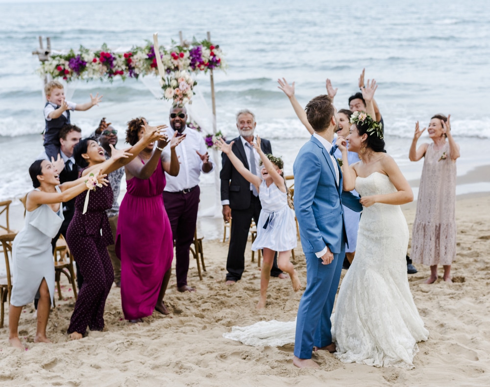A joyful beach wedding scene with guests wearing colorful formal dresses and suits on the sand.