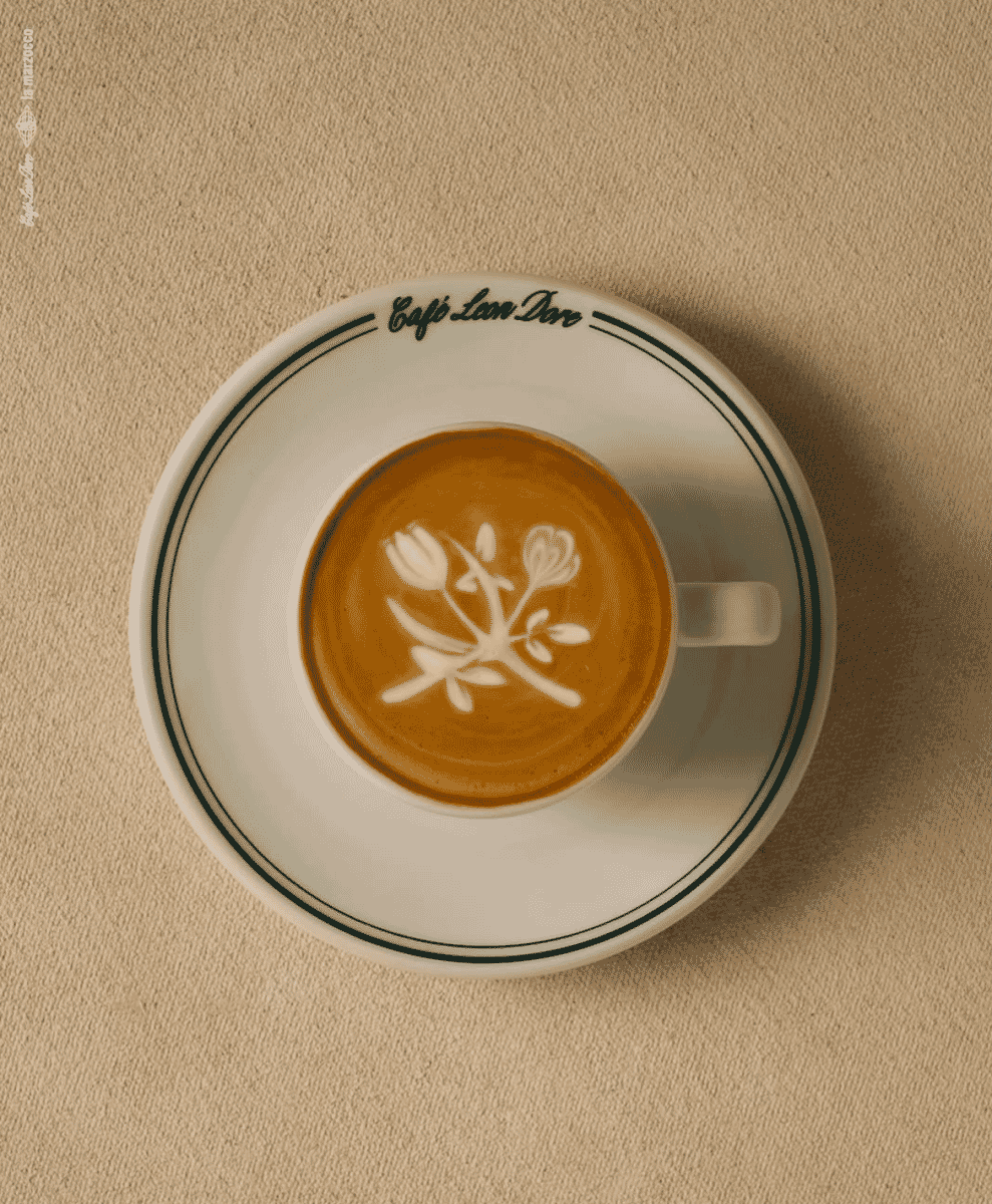 Top-down view of latte art in a Café Leon Dore branded cup on a beige surface.