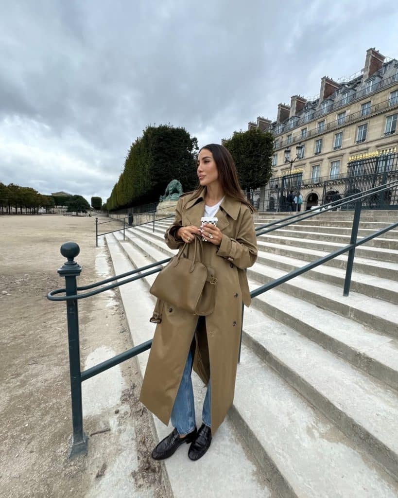 Woman in a beige trench coat and black loafers standing on urban steps.
