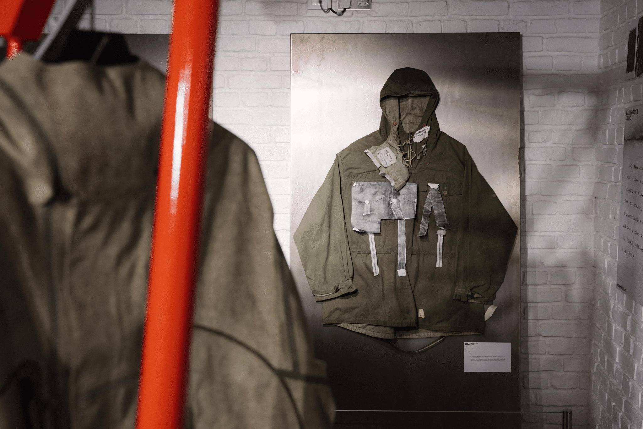 An olive green hooded military-style jacket with attached pouches and straps displayed on a wall panel.