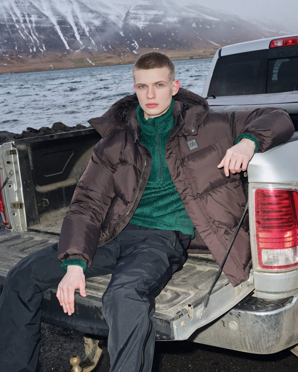 A model in a brown puffer jacket and green turtleneck sitting in a silver pickup truck with mountain scenery.