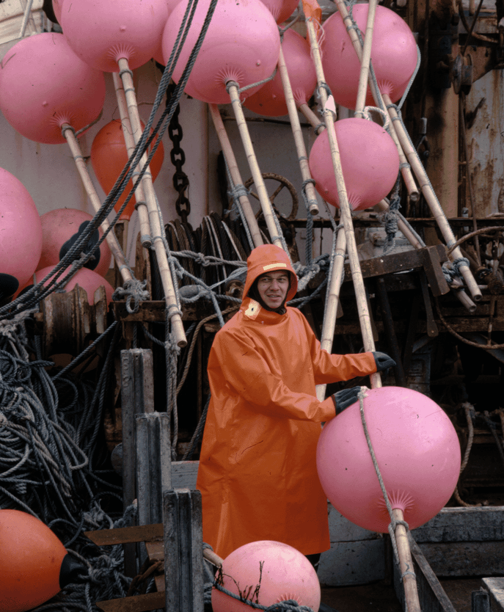 A fisherman in a bright orange waterproof raincoat standing near pink buoys.