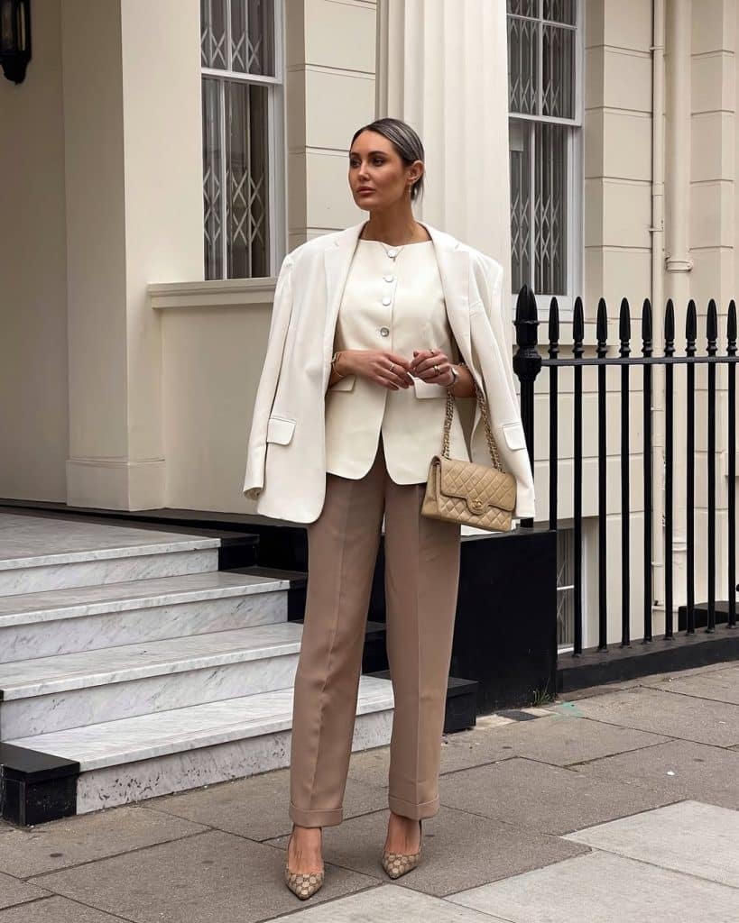 A woman in a cream blazer and taupe trousers carrying a quilted handbag.