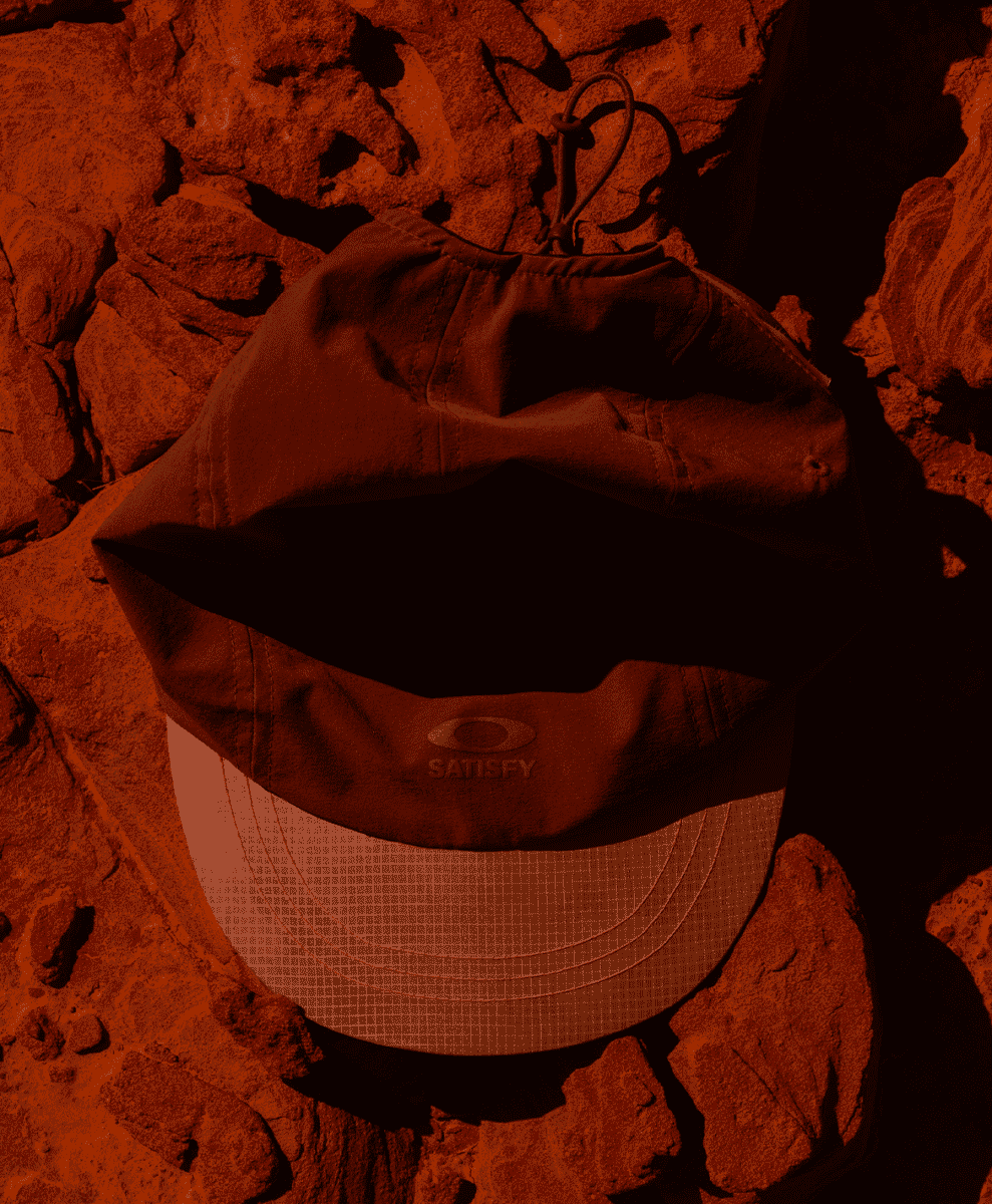 A brown Satisfy x Oakley technical cap resting on jagged reddish-brown rocks.