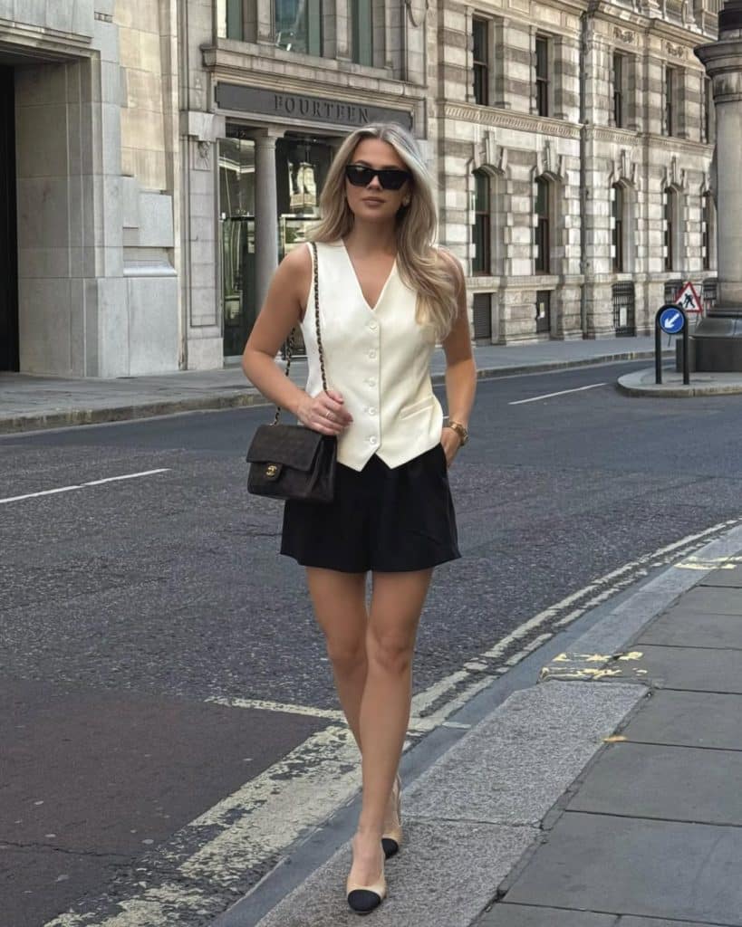 A woman in a cream vest and black shorts carrying a classic Chanel bag on a city street.