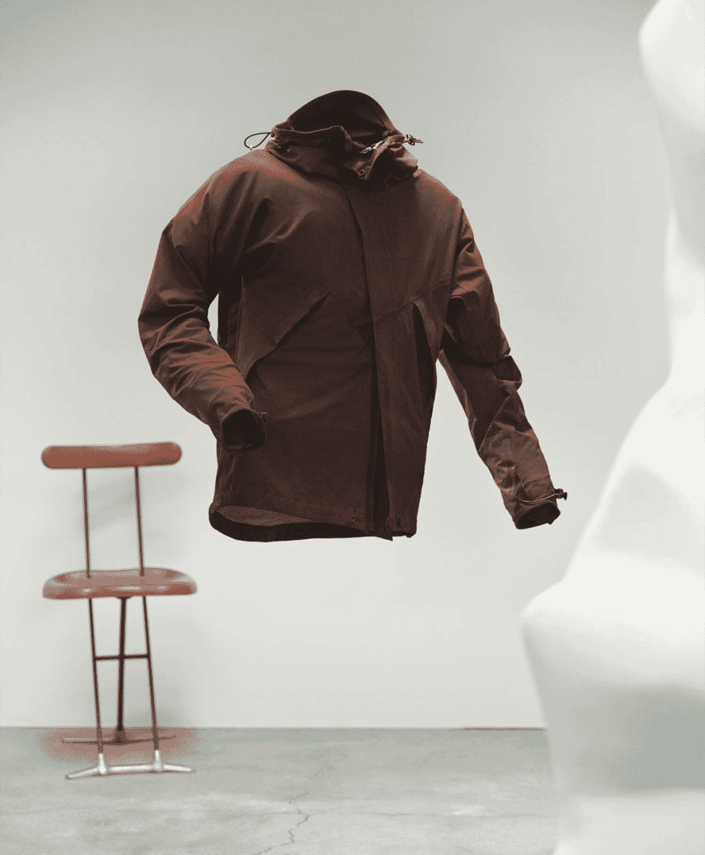 A brown hooded outdoor jacket floating in a studio next to a white mannequin and modern stool.