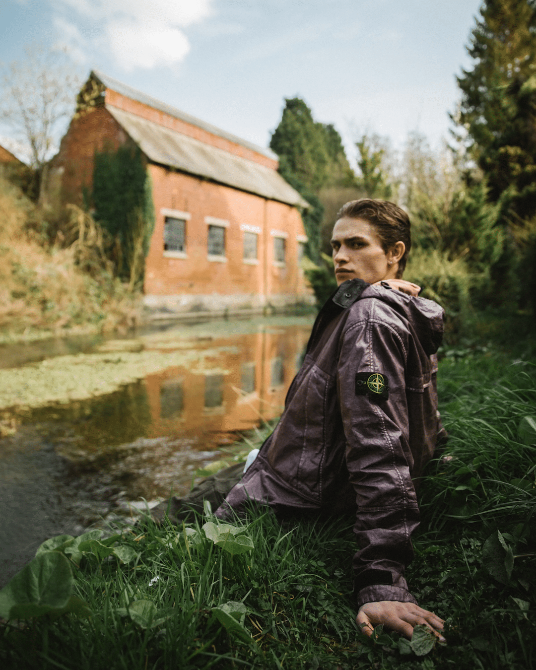 A man in a purple Stone Island coat sitting by a pond near a rustic brick building.