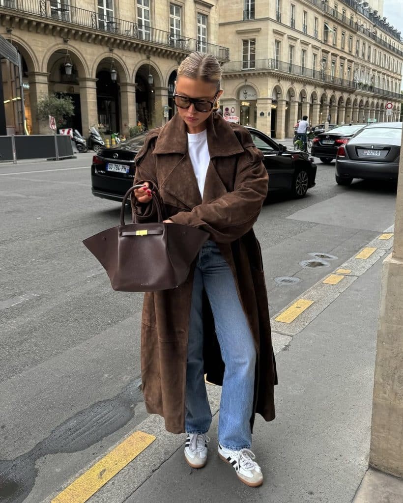 Woman in a brown suede coat and white tee with blue jeans in a city setting.