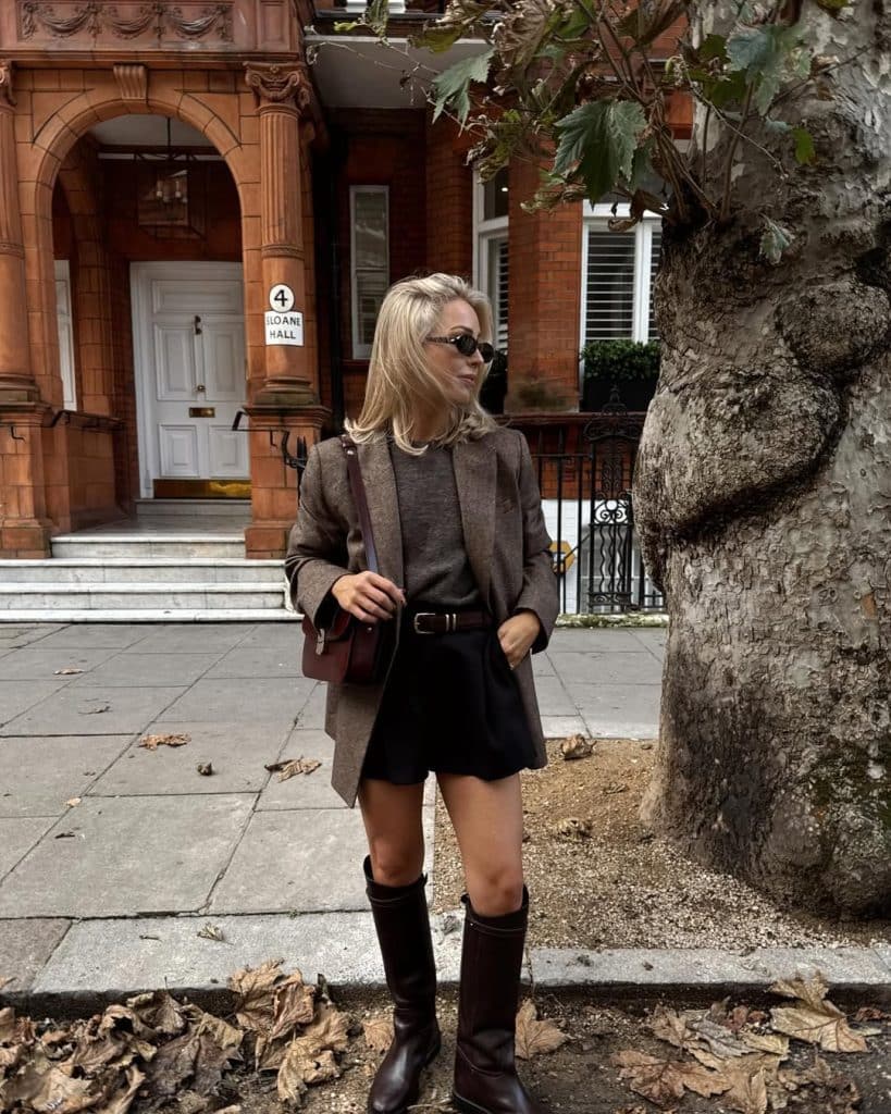 A woman in a brown blazer and black shorts with brown knee-high boots on a city street.