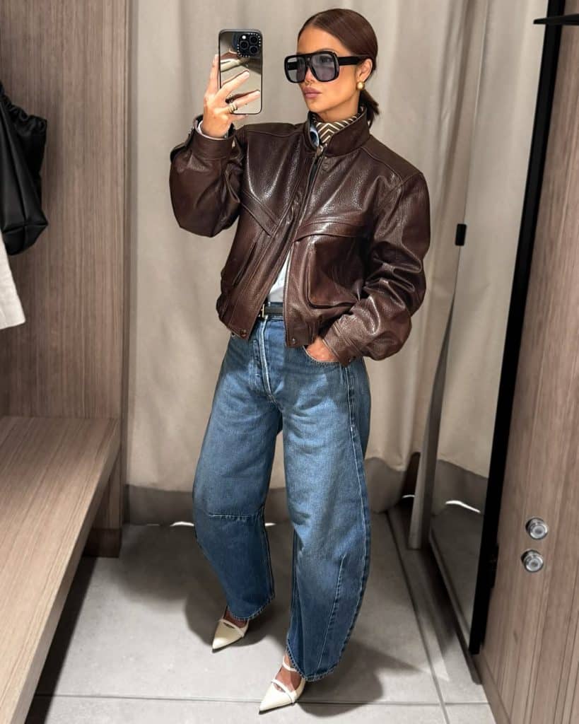 Mirror selfie of a woman in a brown leather bomber jacket and white pointed-toe heels paired with barrel jeans.