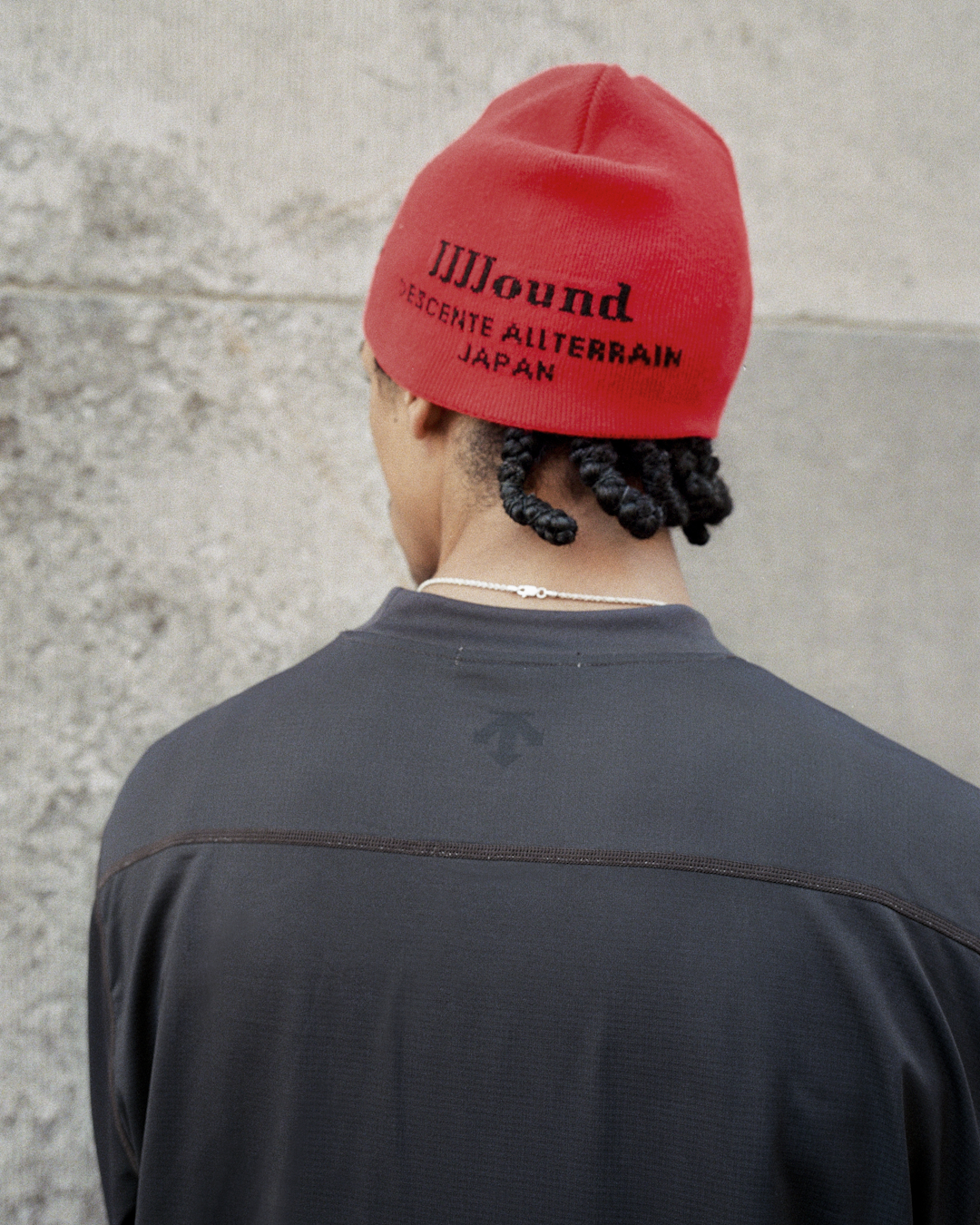 Back view of a person wearing a red knit beanie with 'JJJound DESCENTE ALLTERRAIN JAPAN' text.