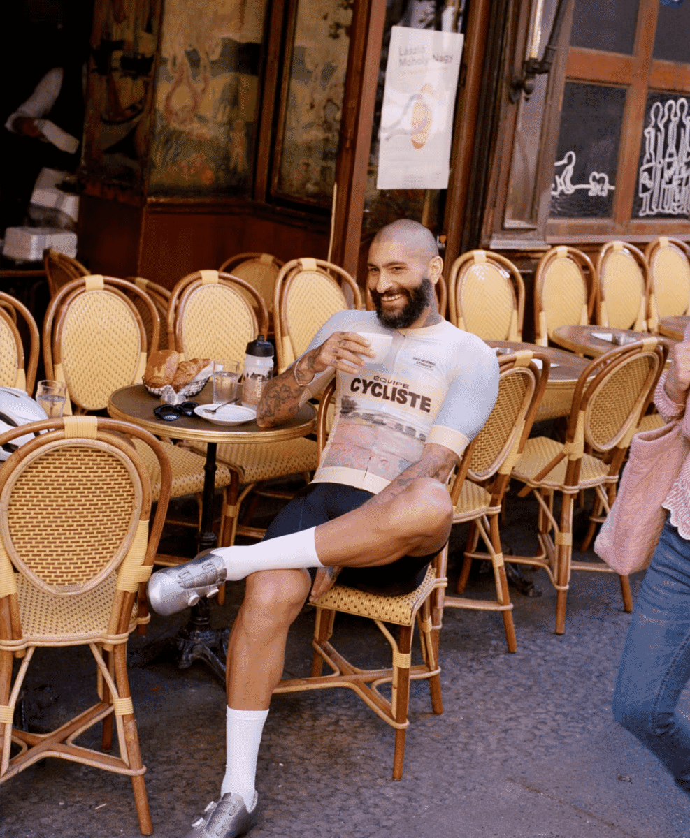 A man in a white cycling jersey sitting at a Parisian café table with a cup of coffee.