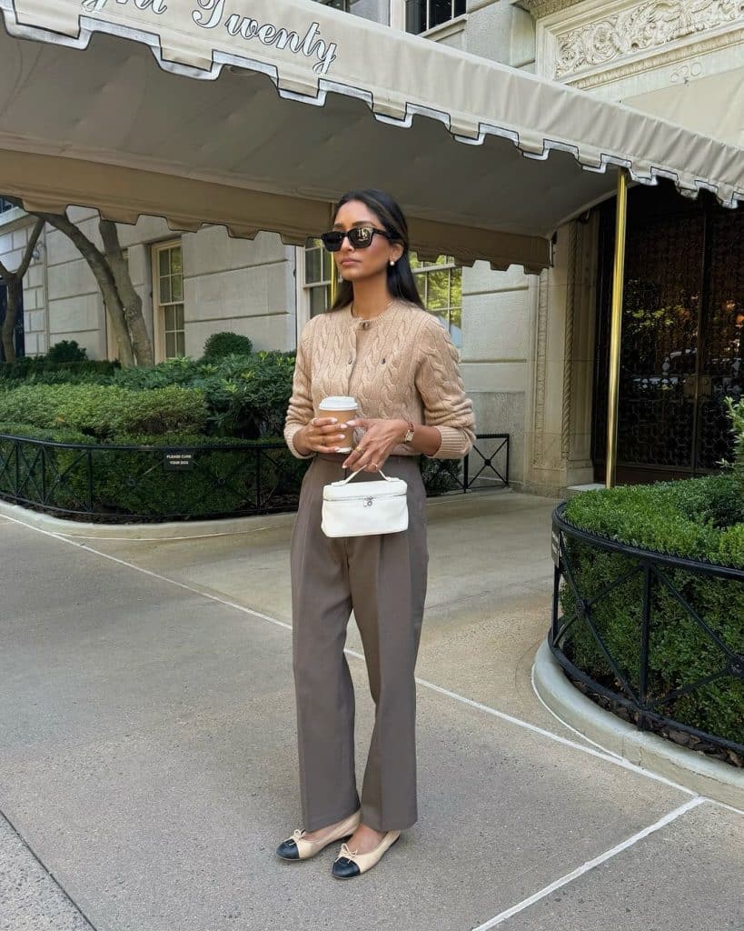 A woman in a beige cable-knit cardigan and taupe wide-leg trousers holding a coffee cup.