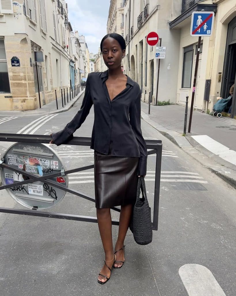 A woman wearing a black silk blouse and a brown leather midi skirt on a city street.