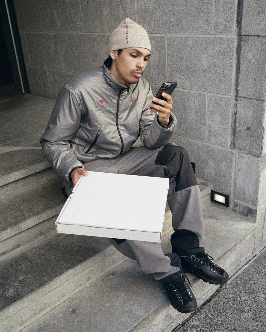 Man in a grey technical jacket and matching beanie sitting on concrete steps while using a phone.