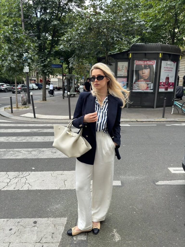 A blonde woman in a navy blazer and cream wide-leg trousers walking on a Parisian street.