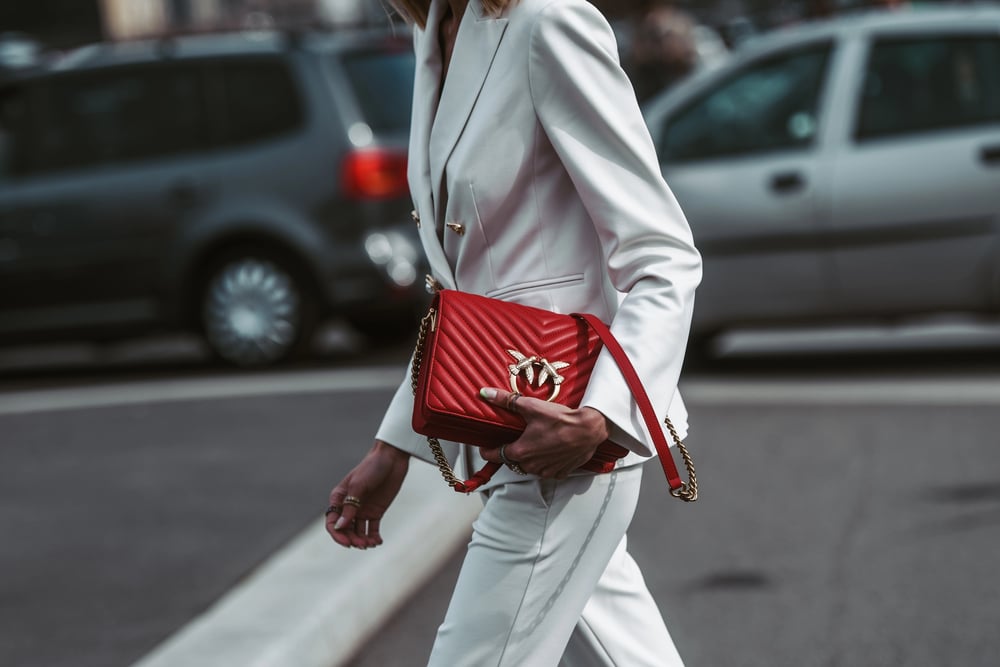 A woman in a white tailored suit carrying a vibrant red quilted handbag on a city street.