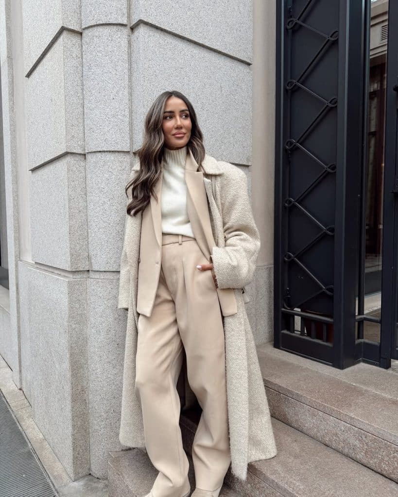 A woman in a layered beige winter outfit featuring a turtleneck, blazer, and textured coat.