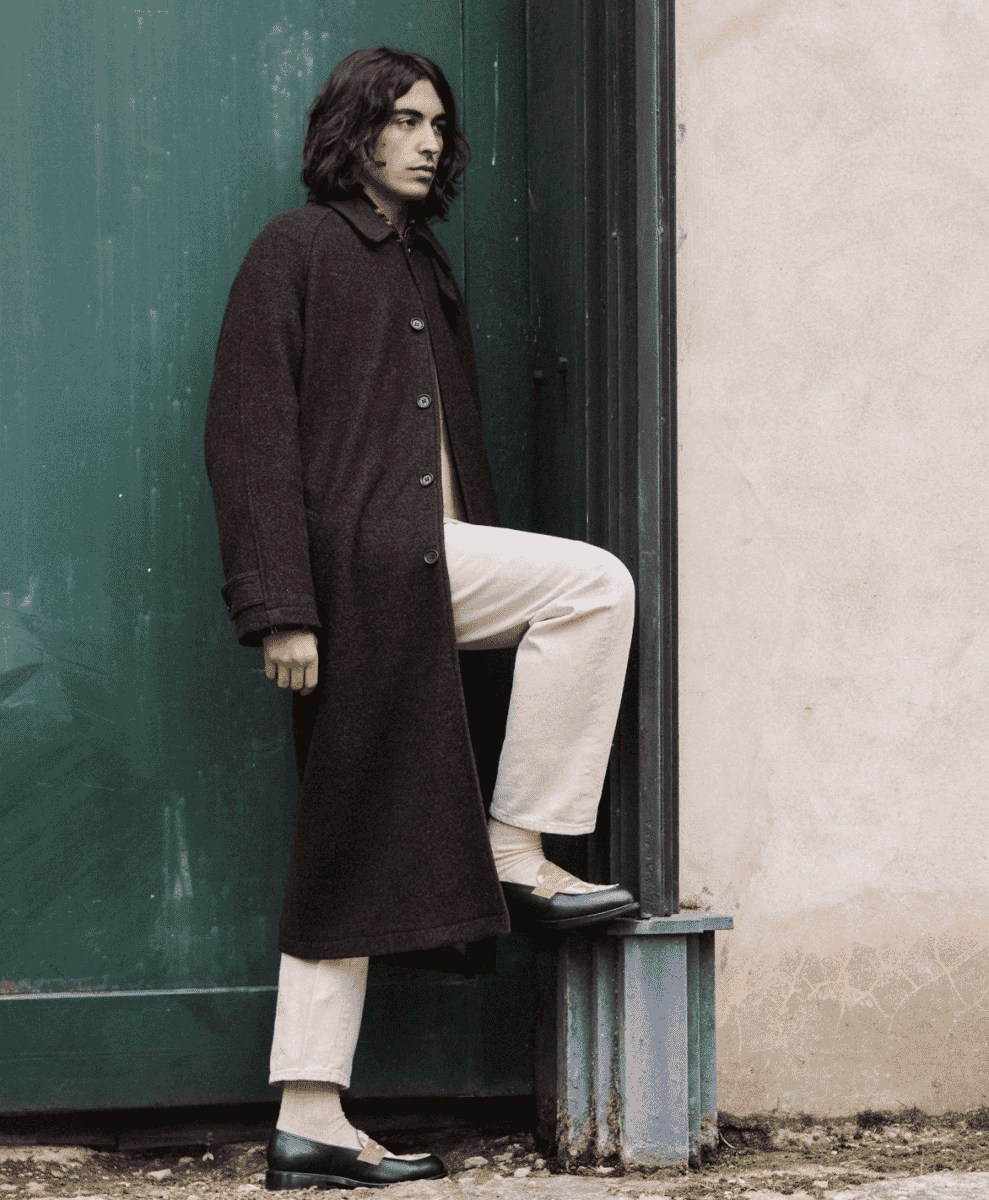 Man in a dark brown wool coat and black loafers standing against a green urban door.