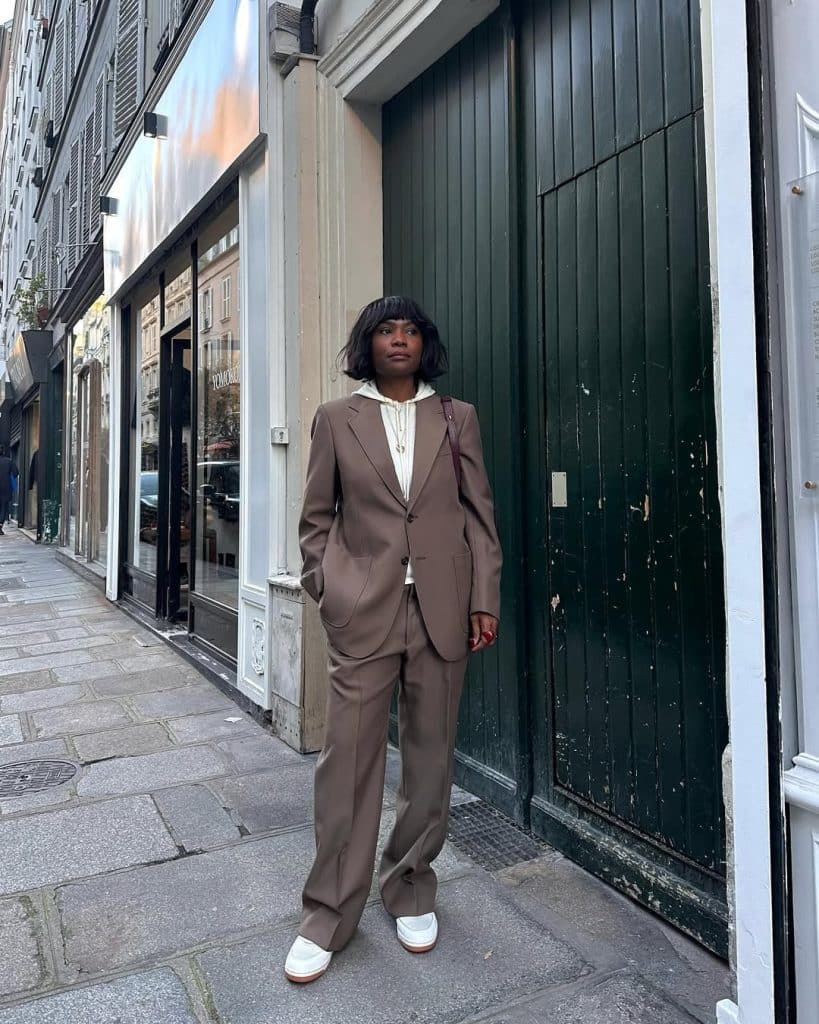 A woman wearing a brown oversized suit with a white hoodie and sneakers.
