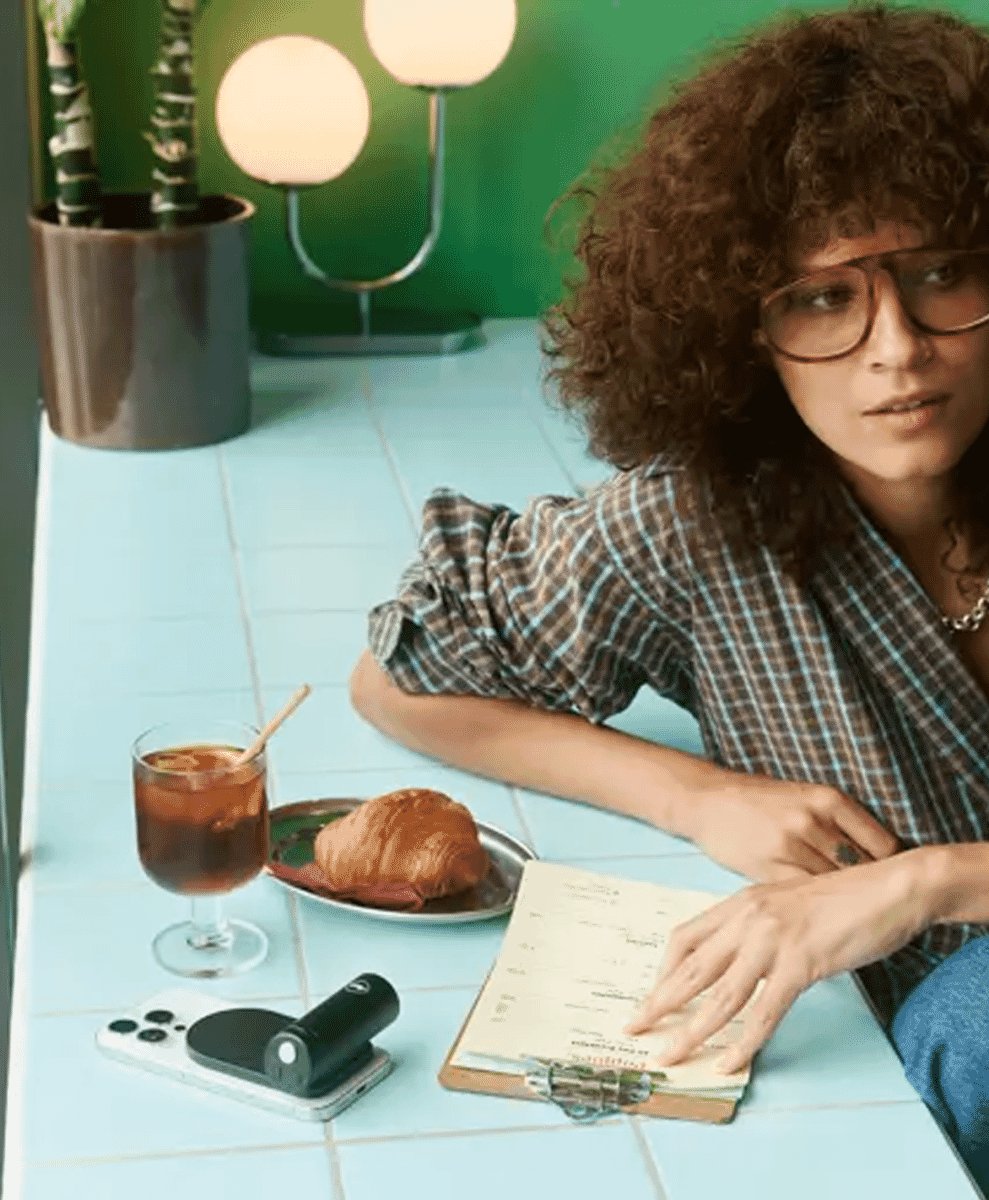 A woman with curly hair in a plaid shirt sitting in a café with a notebook and coffee.