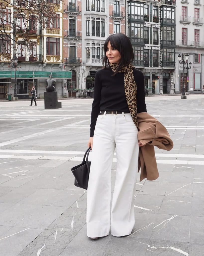 A woman in white wide-leg pants and a black turtleneck accessorized with a leopard-print scarf.