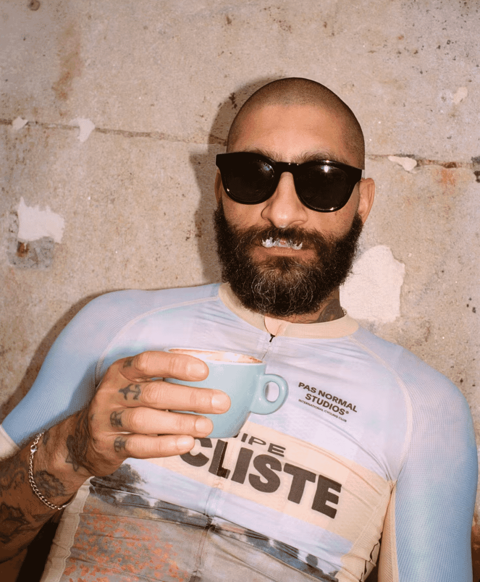 A man in sunglasses and a light blue cycling jersey holding a coffee cup.