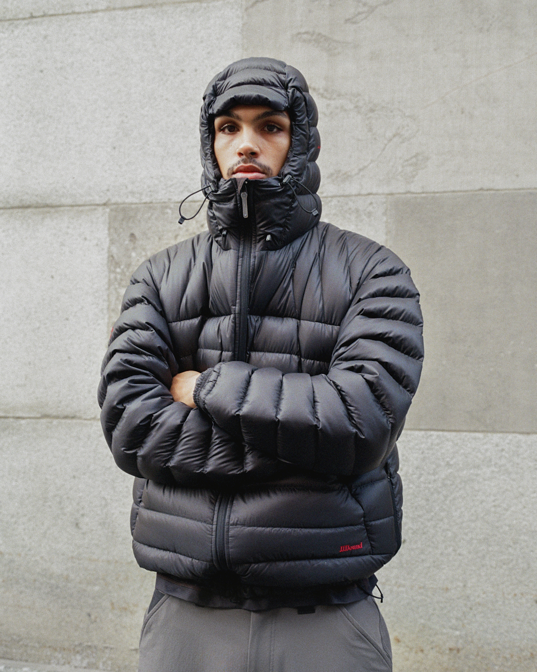 Man in a black hooded puffer jacket standing with arms crossed against a concrete wall.