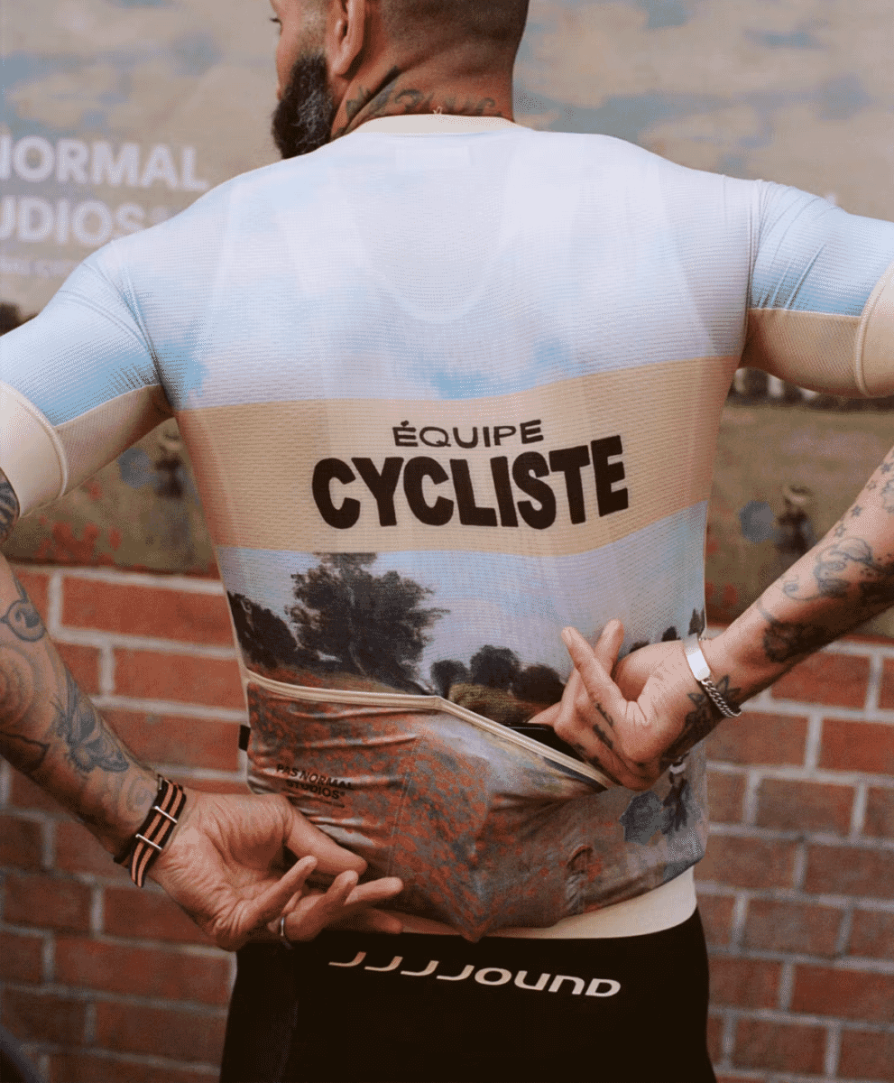 A man wearing a light blue and beige cycling jersey with a landscape print against a brick wall.