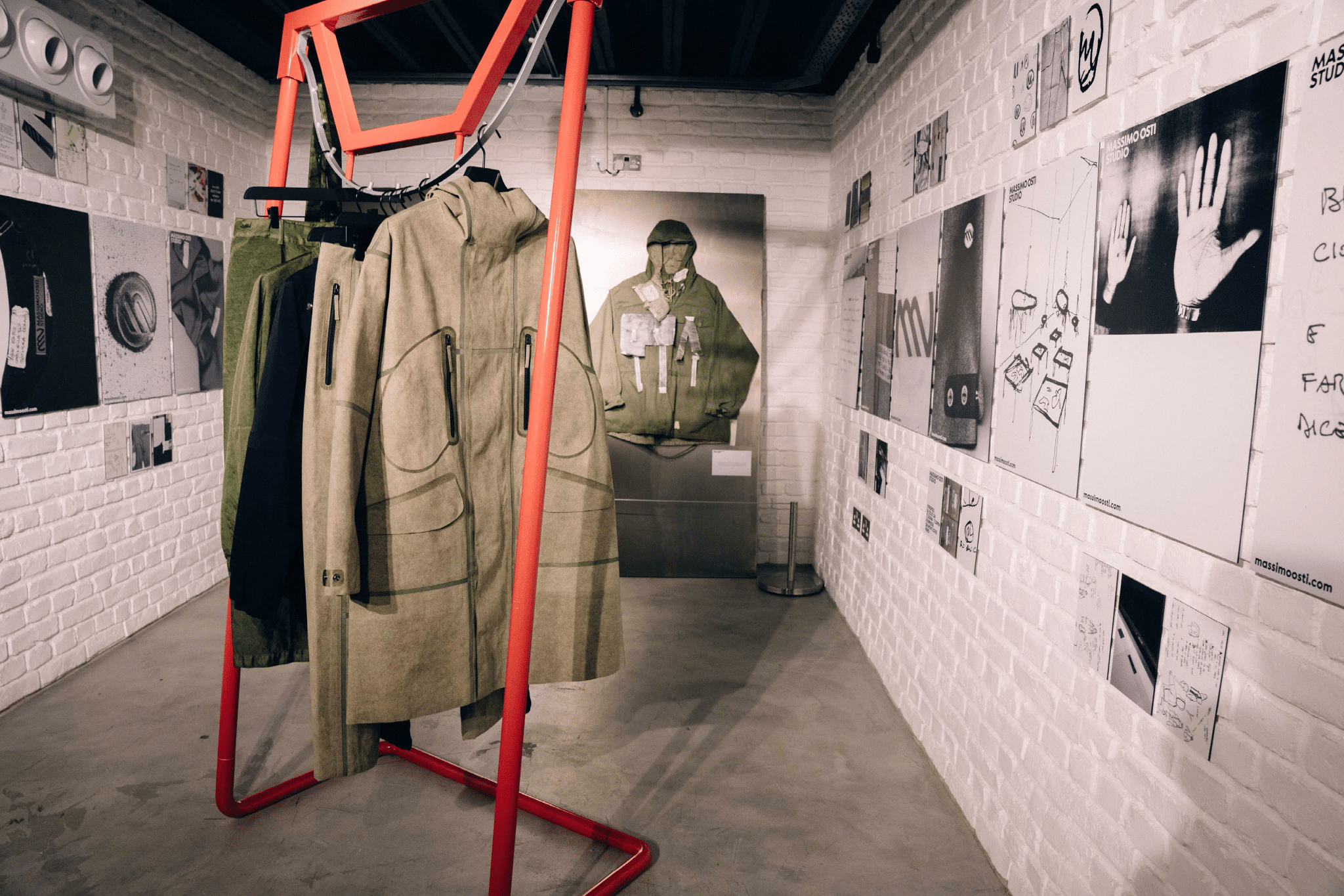 A modern fashion exhibit displays utilitarian coats on a red rack against white brick walls with design posters.
