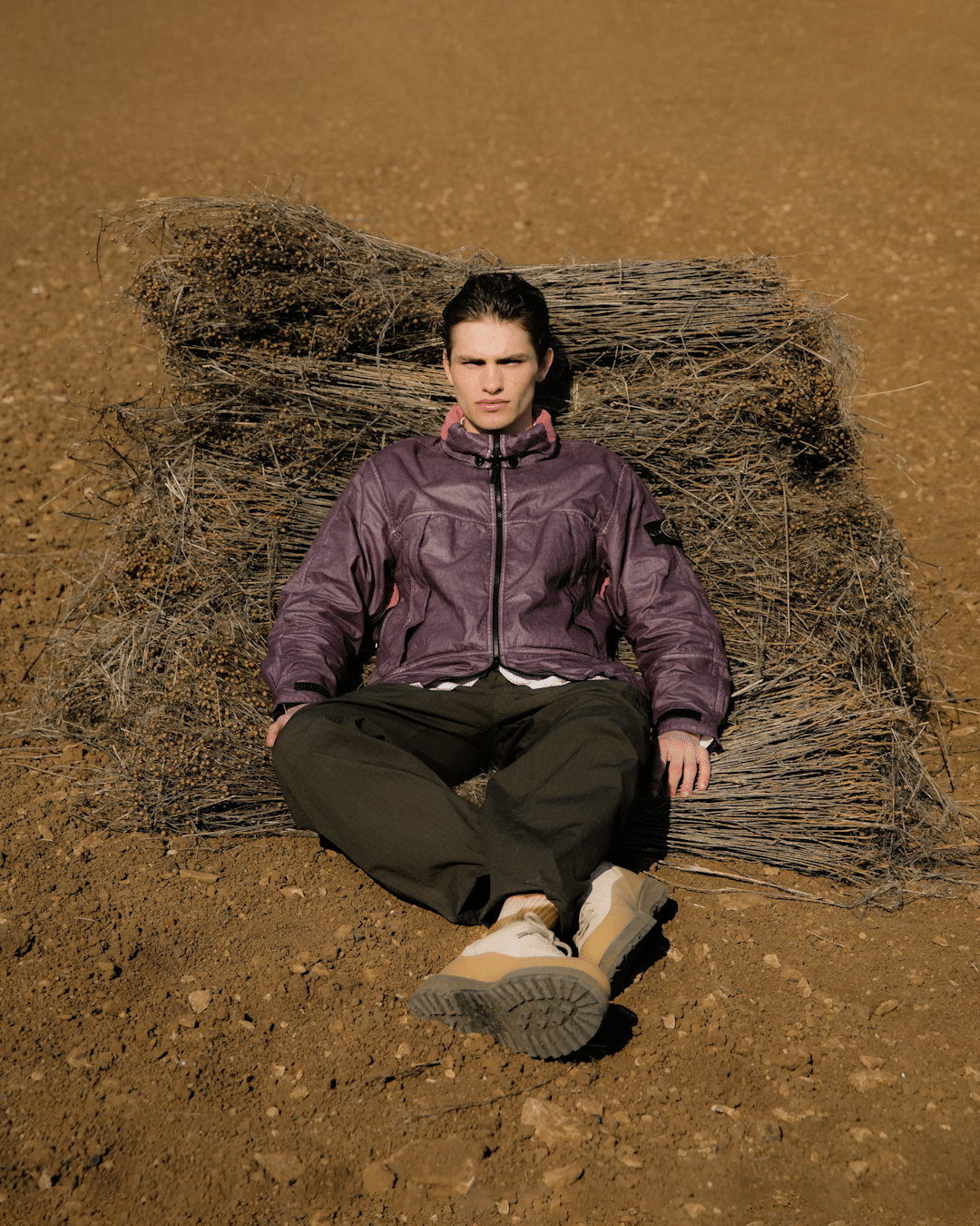 A man sitting on a hay bale in a field wearing a purple Stone Island jacket and rugged boots.