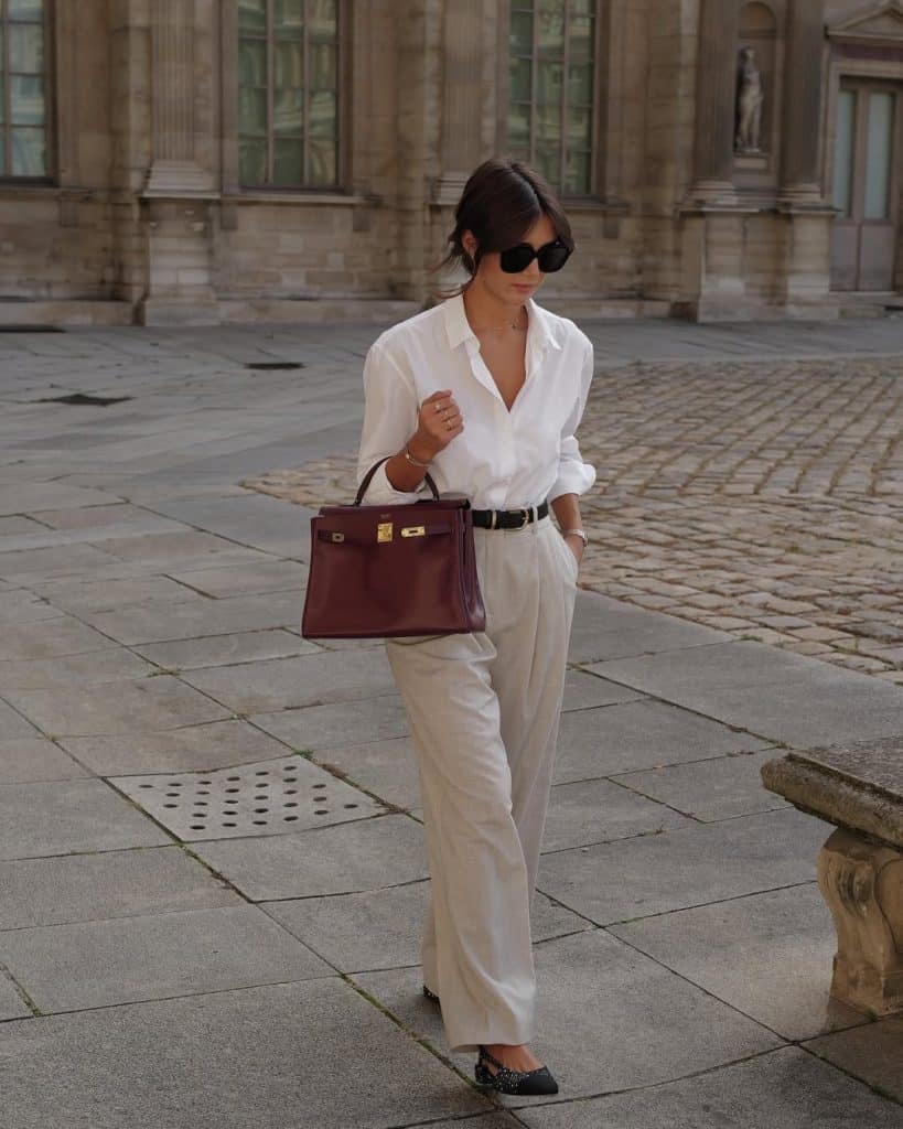 A woman in a white blouse and wide-leg beige trousers carrying a luxury burgundy handbag.